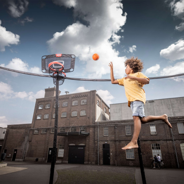 A person jumping on a trampoline, aiming a basketball at the BERG TwinHoop Basketball Hoop.
