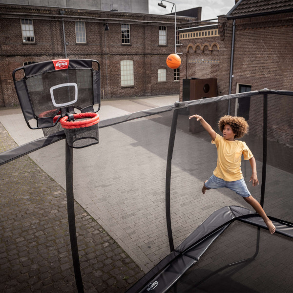 A person shooting a basketball while bouncing on a trampoline with the BERG TwinHoop Basketball Hoop attached.
