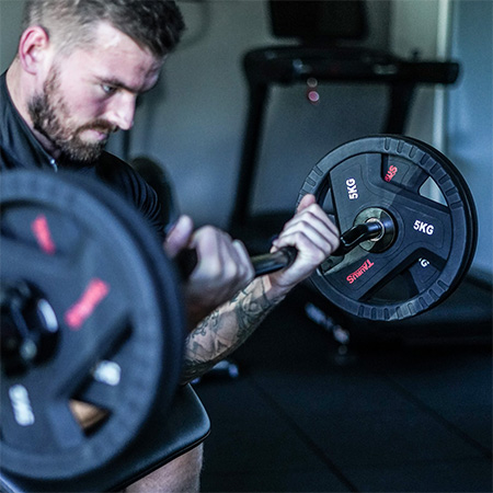 Person performing bicep curls with a barbell loaded with Taurus TPU Olympic Weight Plates in a gym.