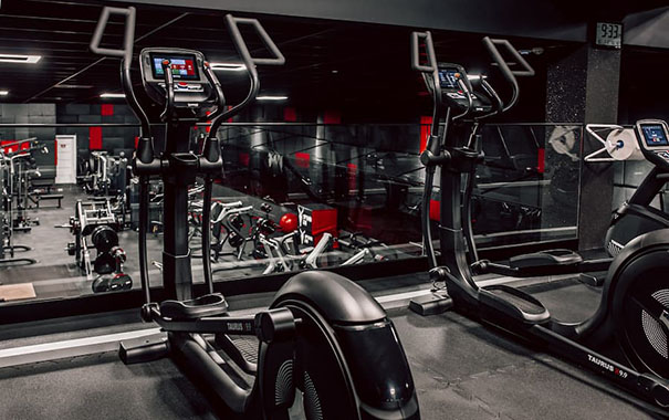 Two elliptical crosstrainers standing in a gym in the first floor. In the background is standing many fitness equipment machines at ground level.