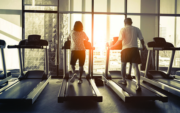 Two people running on treadmills in a gym in front of a windows with sun in the background