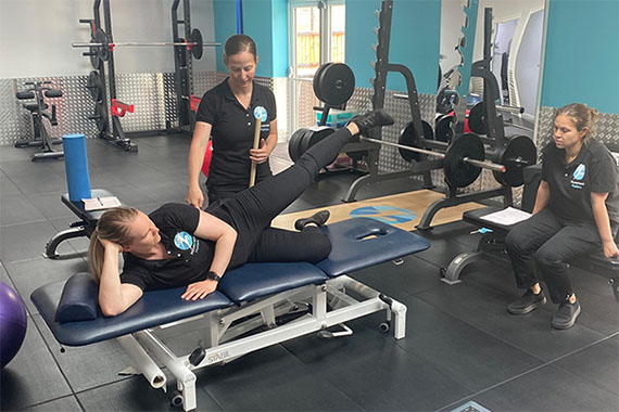 A person is lying on a fitness bench in a gym showing two other persons exercises.