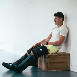 Lifestyle image of the Compex Ayre Compression Boots - Model is sat on a box