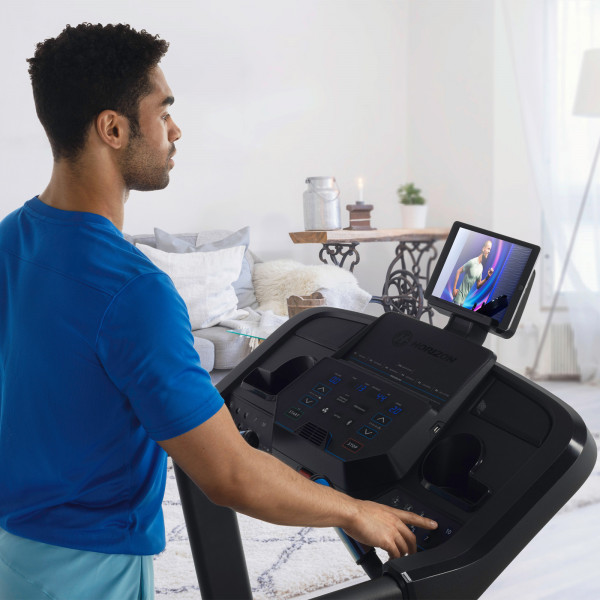 A lifestyle image of a model using the Horizon 5.0AT Treadmill. They are watching a fitness class on a tablet that they have put into the tablet holder. 