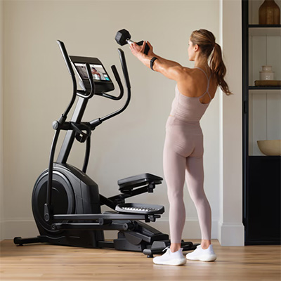 Model performing a dumbbell front raise beside the NordicTrack AirGlide 14i Elliptical in a home workout space   