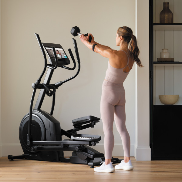 Model performing a dumbbell front raise beside the NordicTrack AirGlide 14i Elliptical in a home workout space