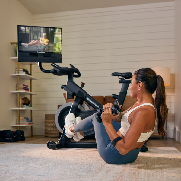 Model performing a core workout with dumbbells beside the NordicTrack X24 Bike with workout on screen