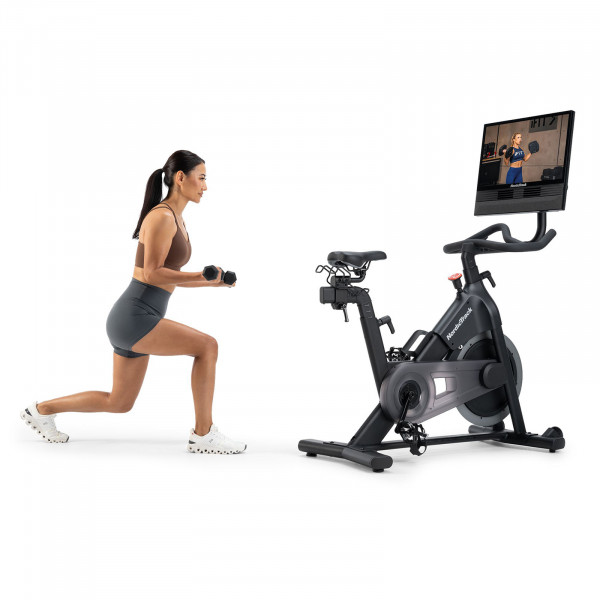 Model performing dumbbell lunges next to NordicTrack 24 Studio Exercise Bike with trainer-led session on screen