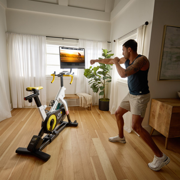 Person stretching near the NordicTrack Tour de France Indoor Bike with the screen displaying a cycling workout.

