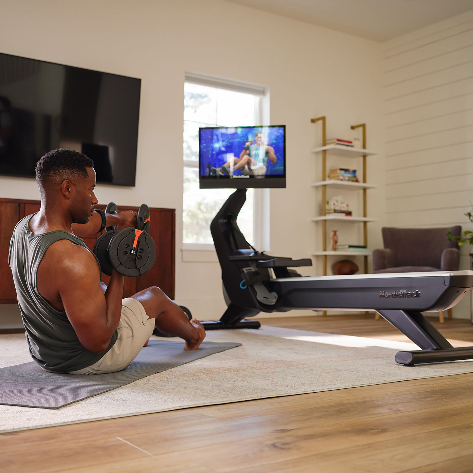 Model performing seated dumbbell curls beside NordicTrack RW900 Rower with workout on screen 