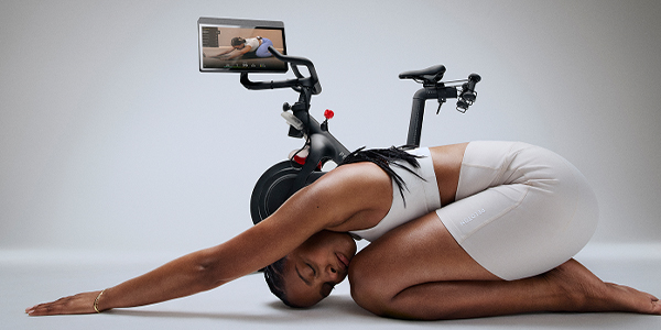 Lifestyle banner for the Peloton Cross Training Bike, with a model doing a stretching yoga pose, matching the position on the swivelled screen on the Bike.