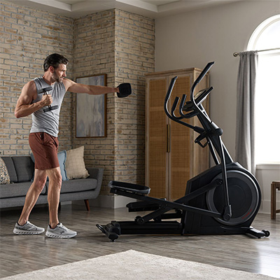 Model doing a dumbbell punch exercise beside the ProForm Carbon EL Elliptical at home  