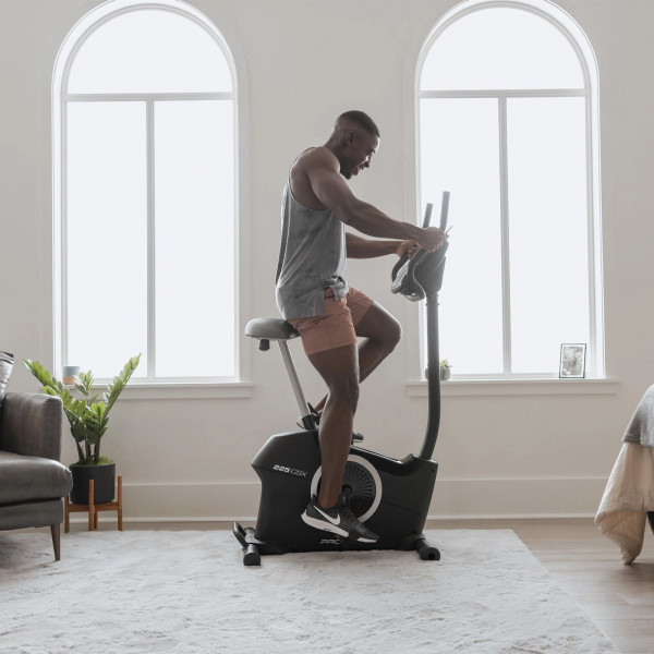 Lifestyle image of the ProForm 225 CSX Upright Exercise Bike showing a model riding the bike from the side in a home setting