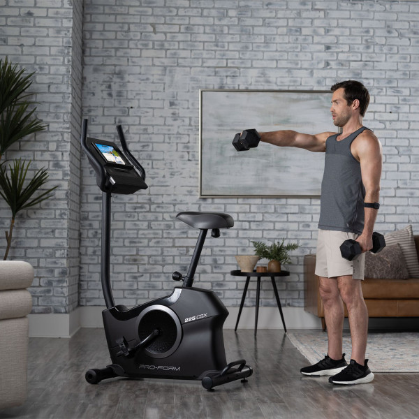 Model performing a dumbbell front raise beside the ProForm 225 CSX Upright Exercise Bike