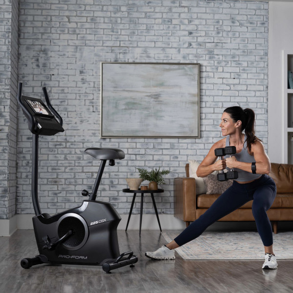 Model performing a side lunge with dumbbells behind the ProForm 225 CSX Upright Exercise Bike