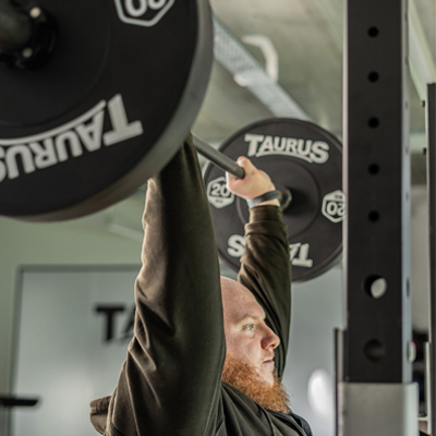 A close-up of a model performing a shoulder press with the Taurus 20 kg Olympic barbell.